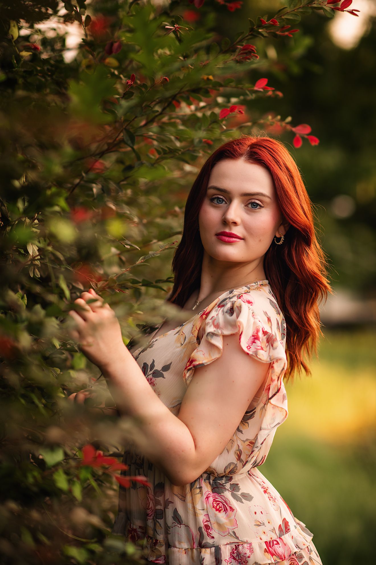 Young woman with vibrant red hair in floral dress posing beside blooming red flowers in a lush garden.