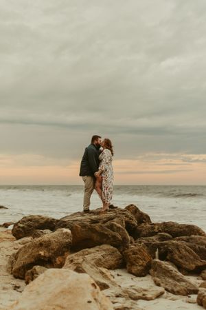 Couple embraces on rocky coastline during dramatic cloudy sunset.
