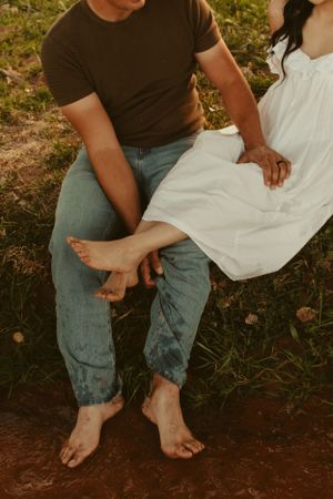 Intimate barefoot couple sitting together on dirt ground in casual attire during golden hour.