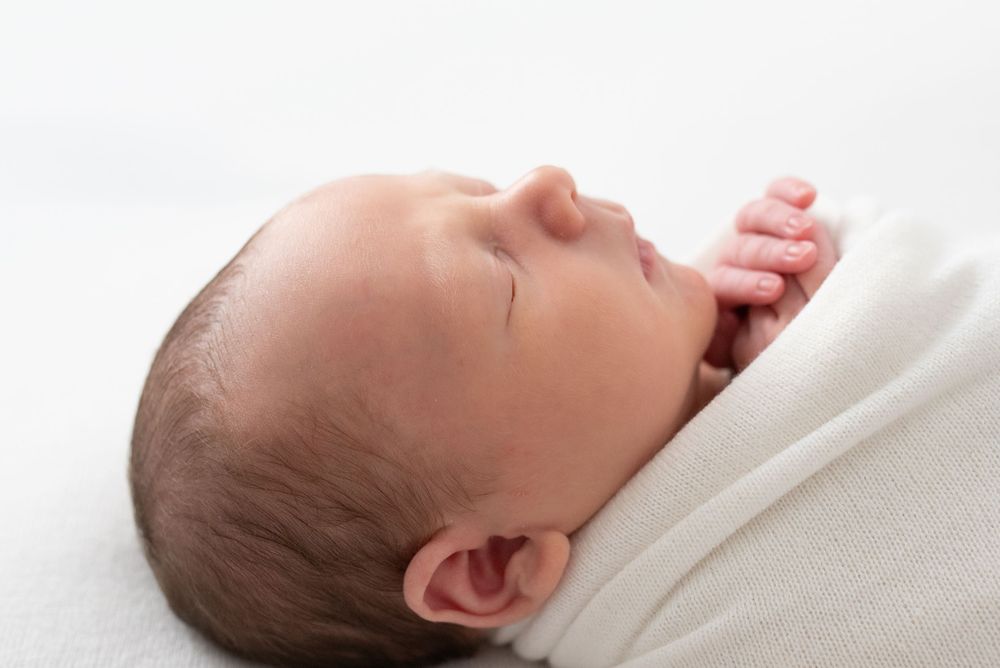 Peaceful newborn baby swaddled in white blanket sleeping, close-up profile view of infant with dark hair.