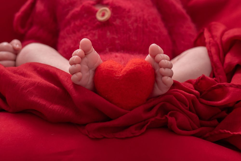 Close-up of newborn baby feet holding a red heart between them, wrapped in soft red blanket.