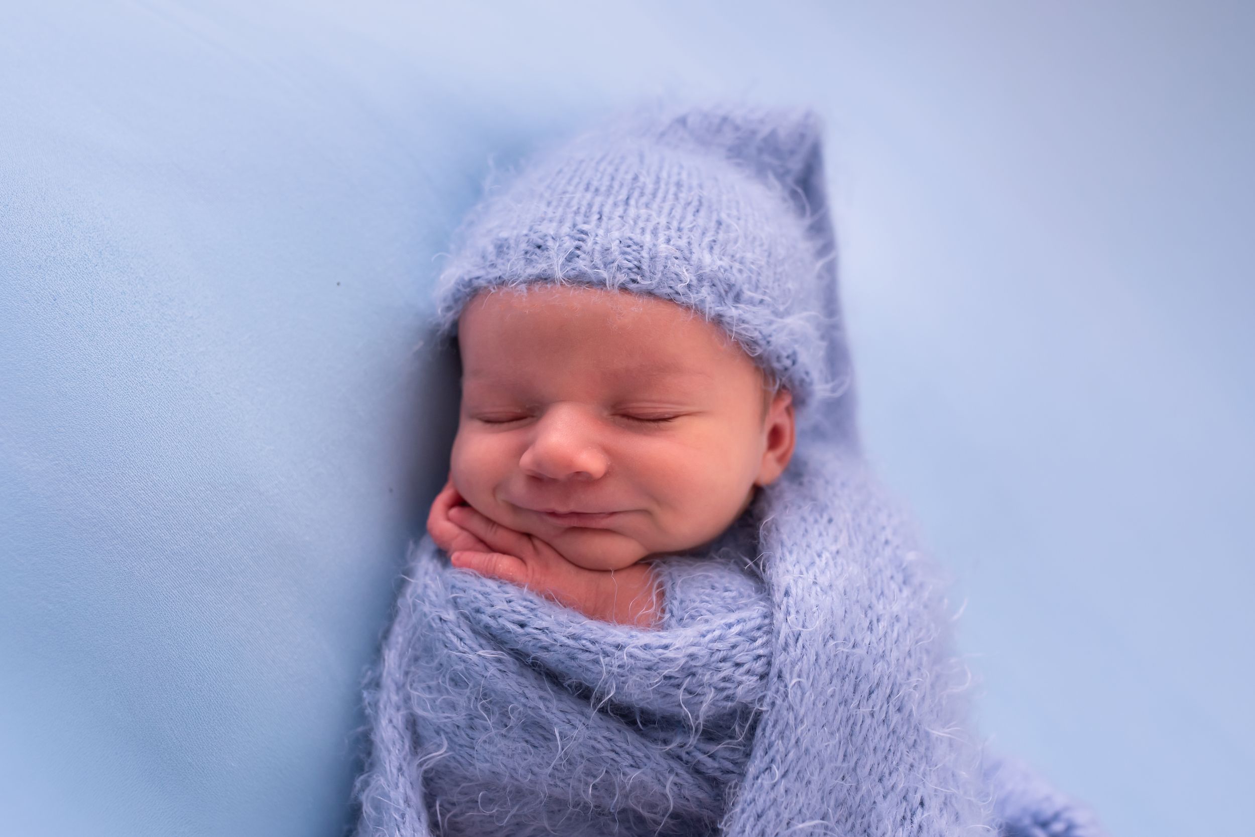Peaceful newborn baby sleeping wrapped in soft blue knit blanket and matching hat on light blue background.