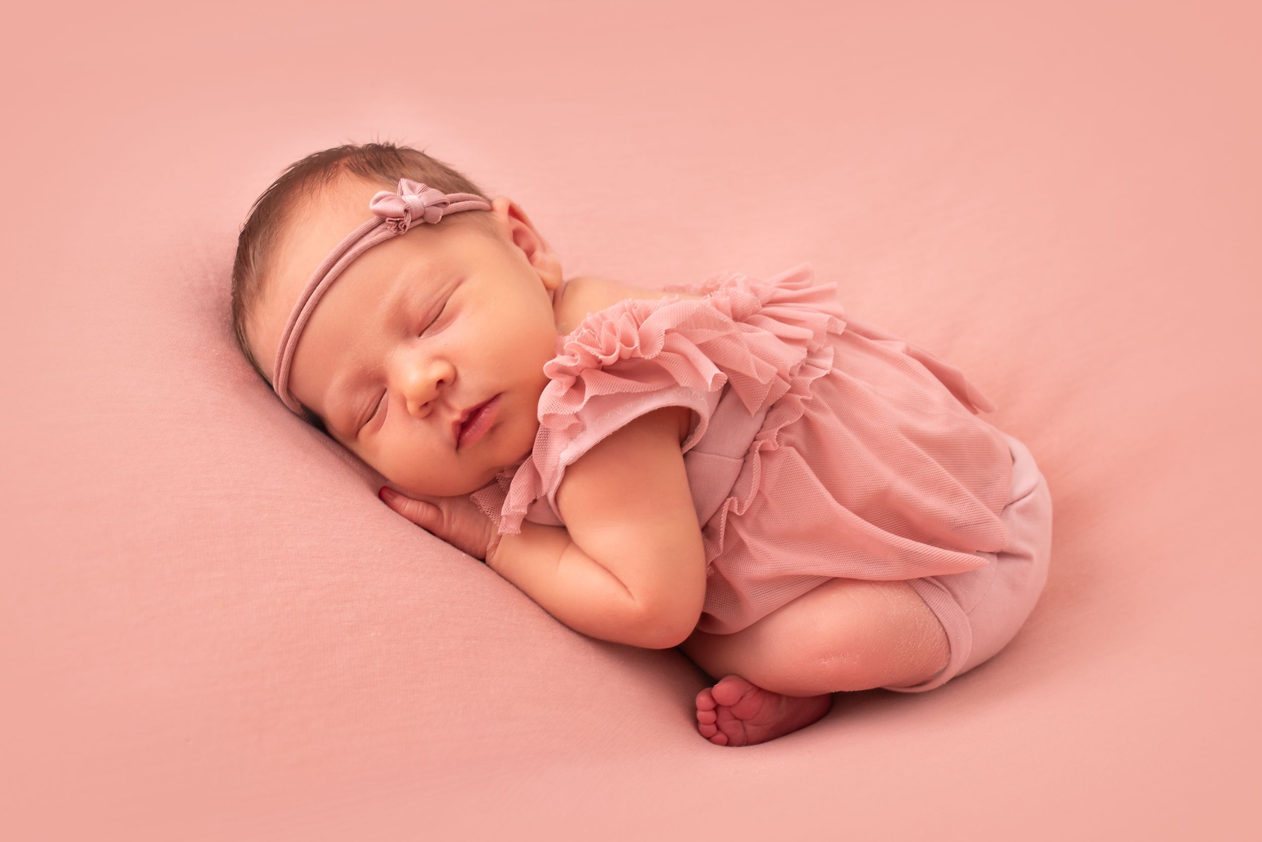 Sleeping newborn baby girl in pink romper and floral headband on soft pink background.