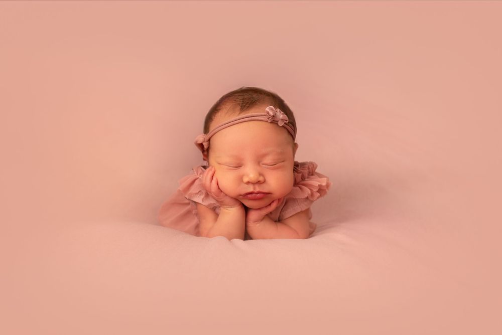 Newborn baby girl sleeping peacefully in froggy pose wearing pink outfit and floral headband on pink backdrop.
