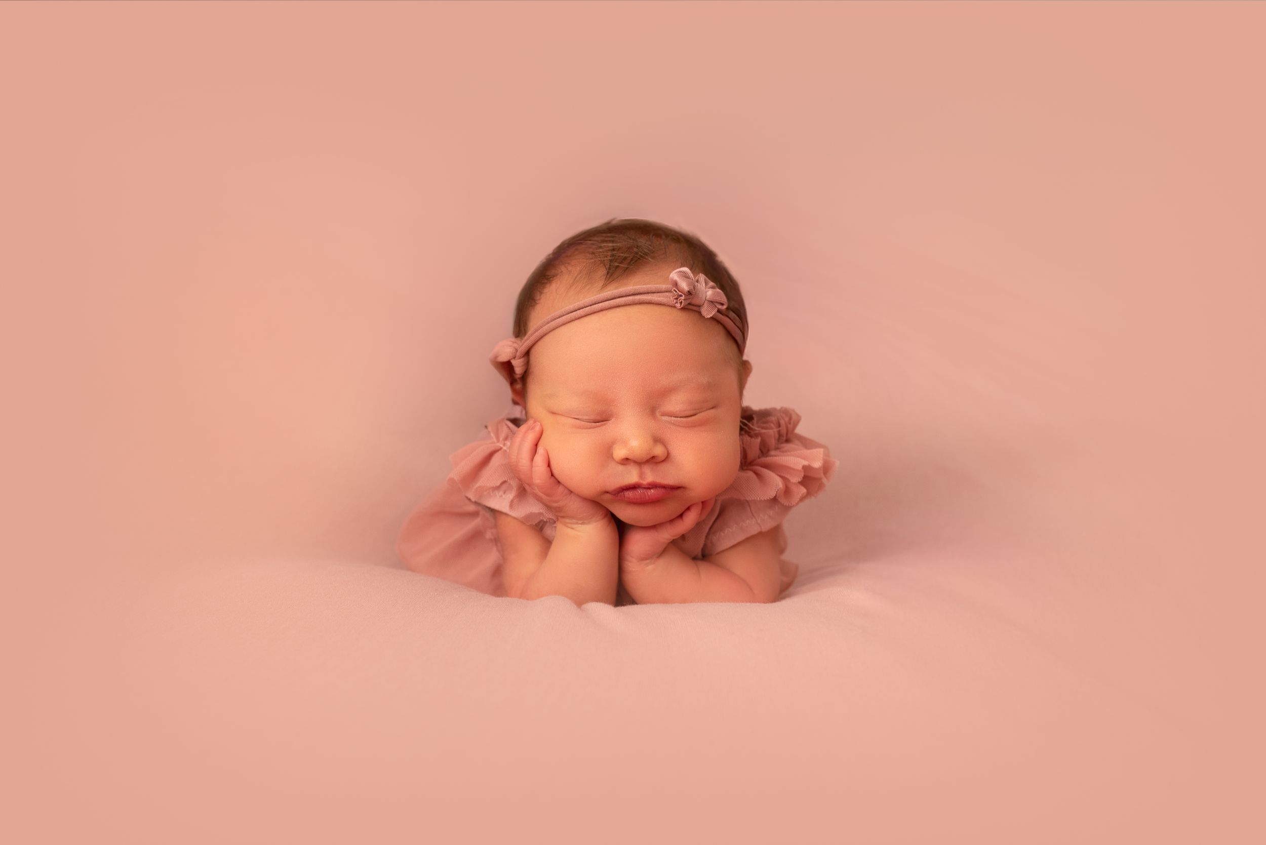 Newborn baby girl sleeping peacefully in froggy pose wearing pink outfit and floral headband on pink backdrop.
