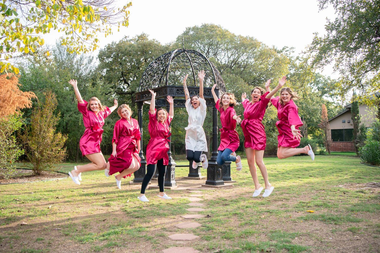 Bridesmaids in matching pink satin robes jump joyfully on a sunny lawn beside a garden gazebo while a bride in white laughs in the center.