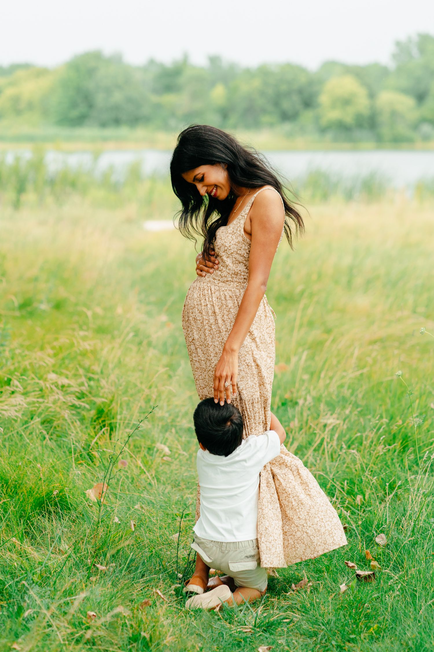 Smiling pregnant woman in a floral dress rests her hand on her belly as her toddler hugs her legs in a grassy lakeside meadow.