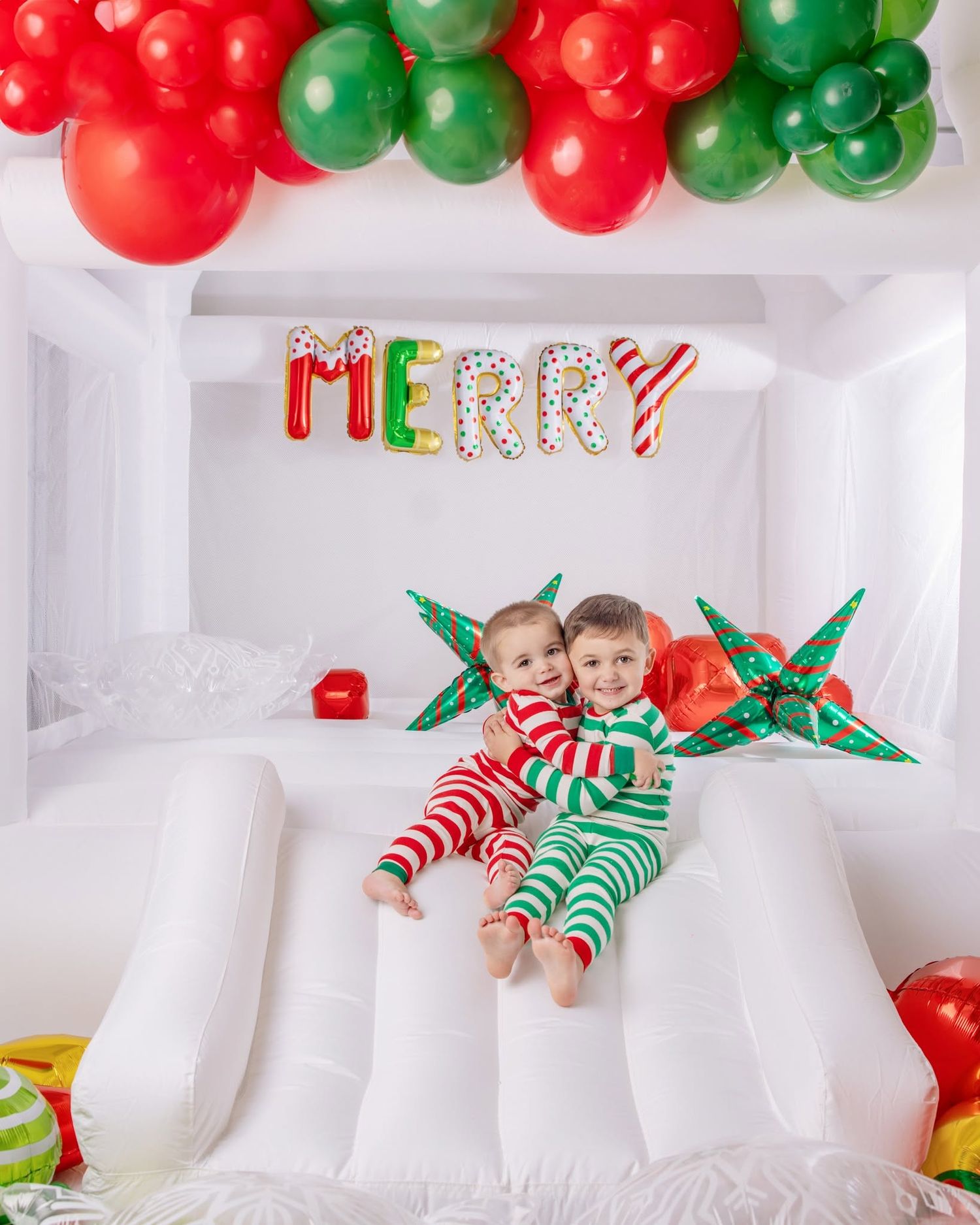 Two children in red and green striped Christmas pajamas sit on a white bounce house slide with a MERRY balloon banner and balloon garland.