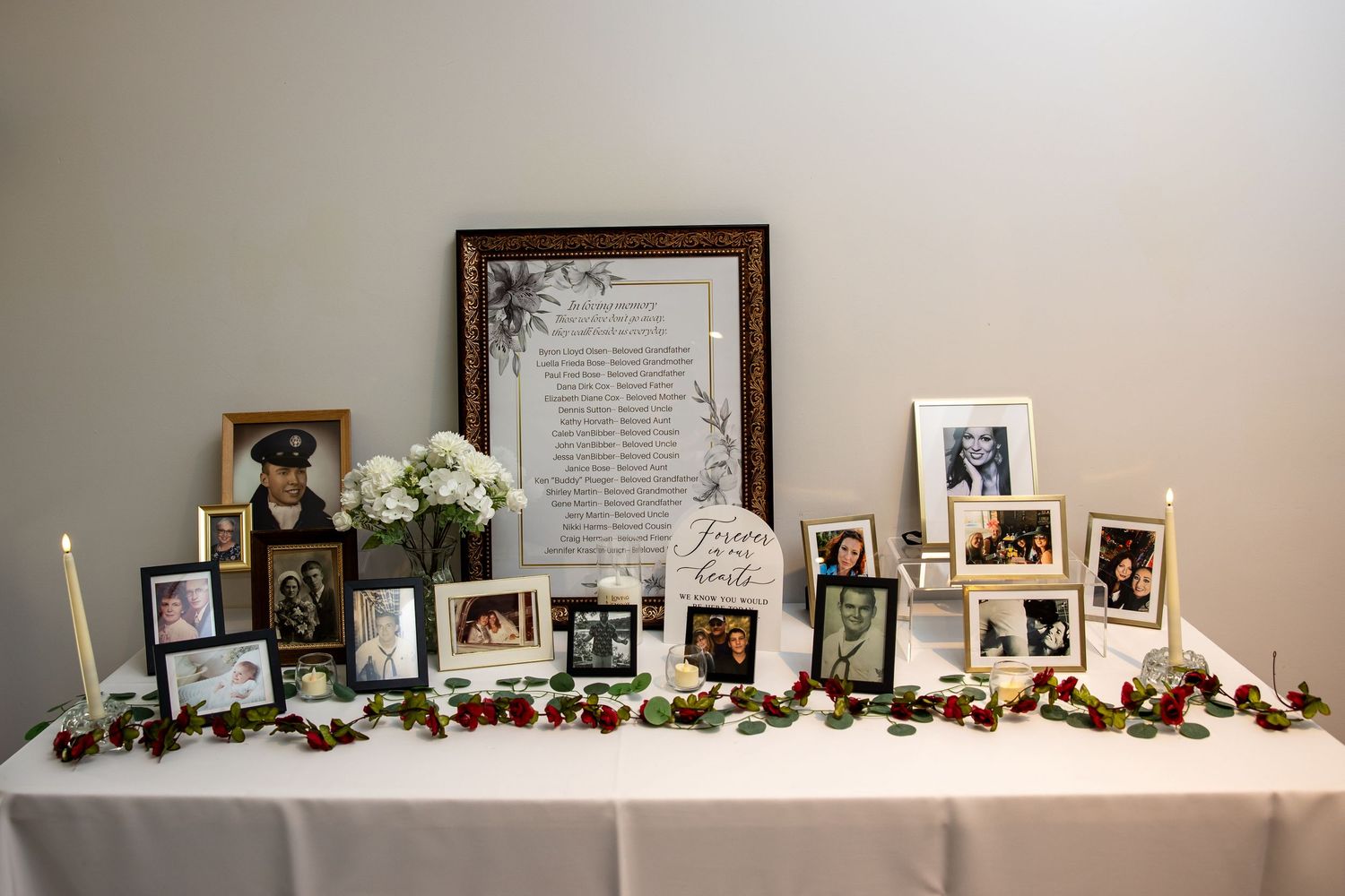 Memorial tribute table with framed family photos, white flowers, lit candles, a framed poem, and a red rose garland on a white tablecloth.