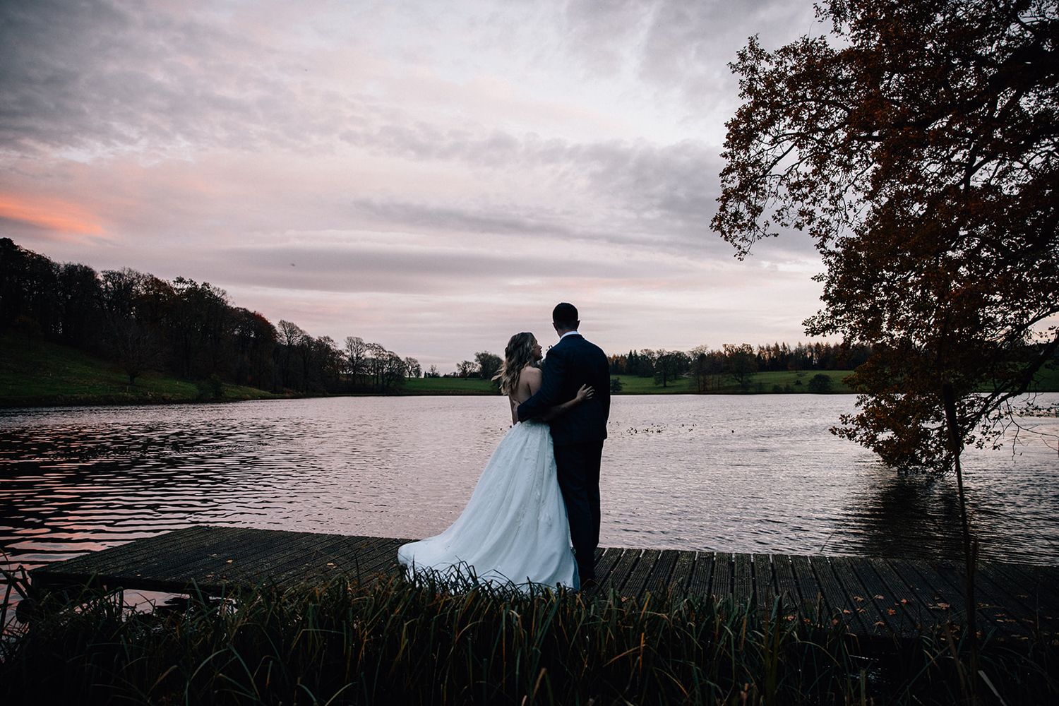 Bride in a white gown and groom in a dark suit embrace on a wooden lake dock at sunset with a pink and purple sky.