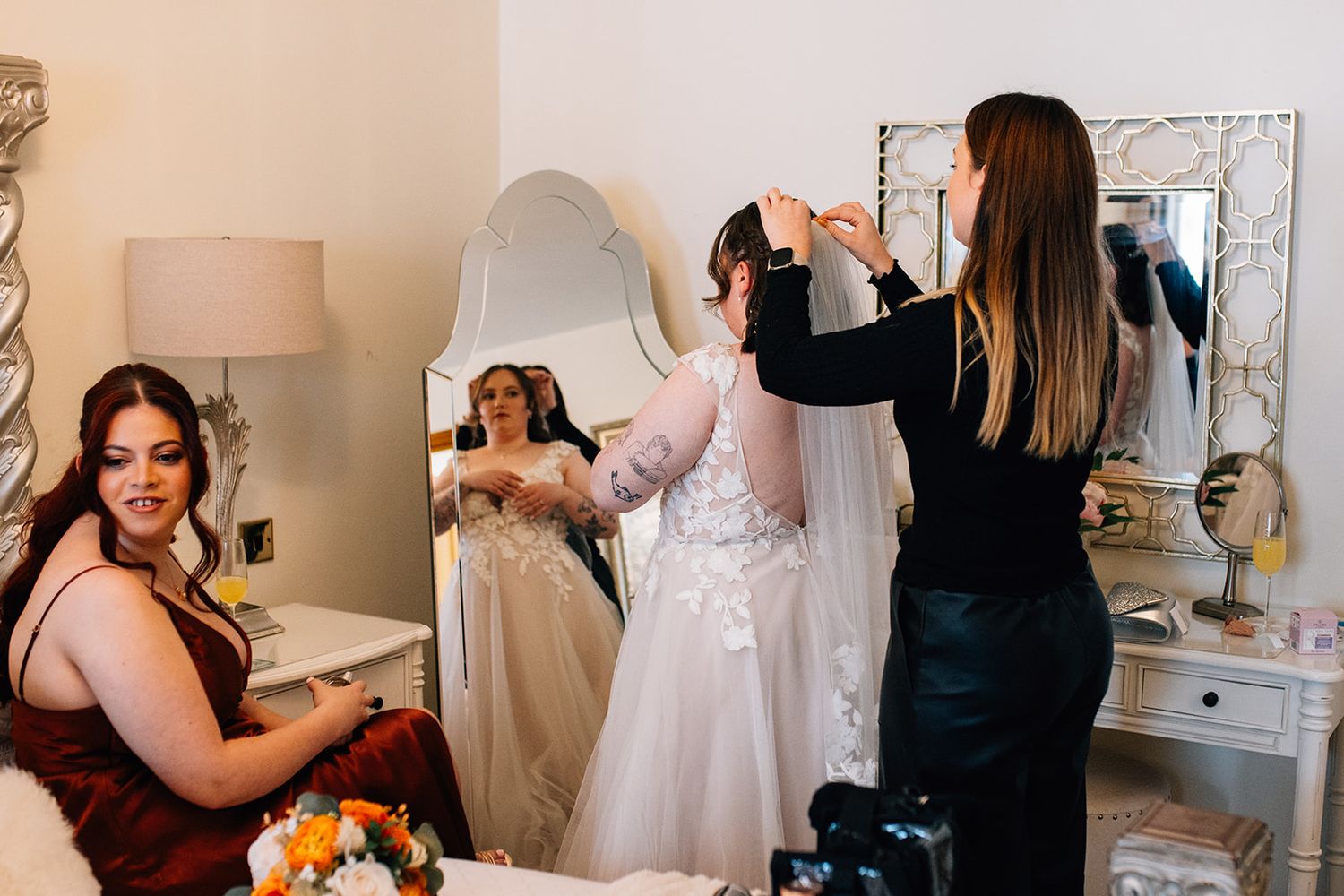 Hair stylist pins a bride's veil in a bright dressing room while a bridesmaid in a burgundy dress watches and a flower bouquet rests nearby.