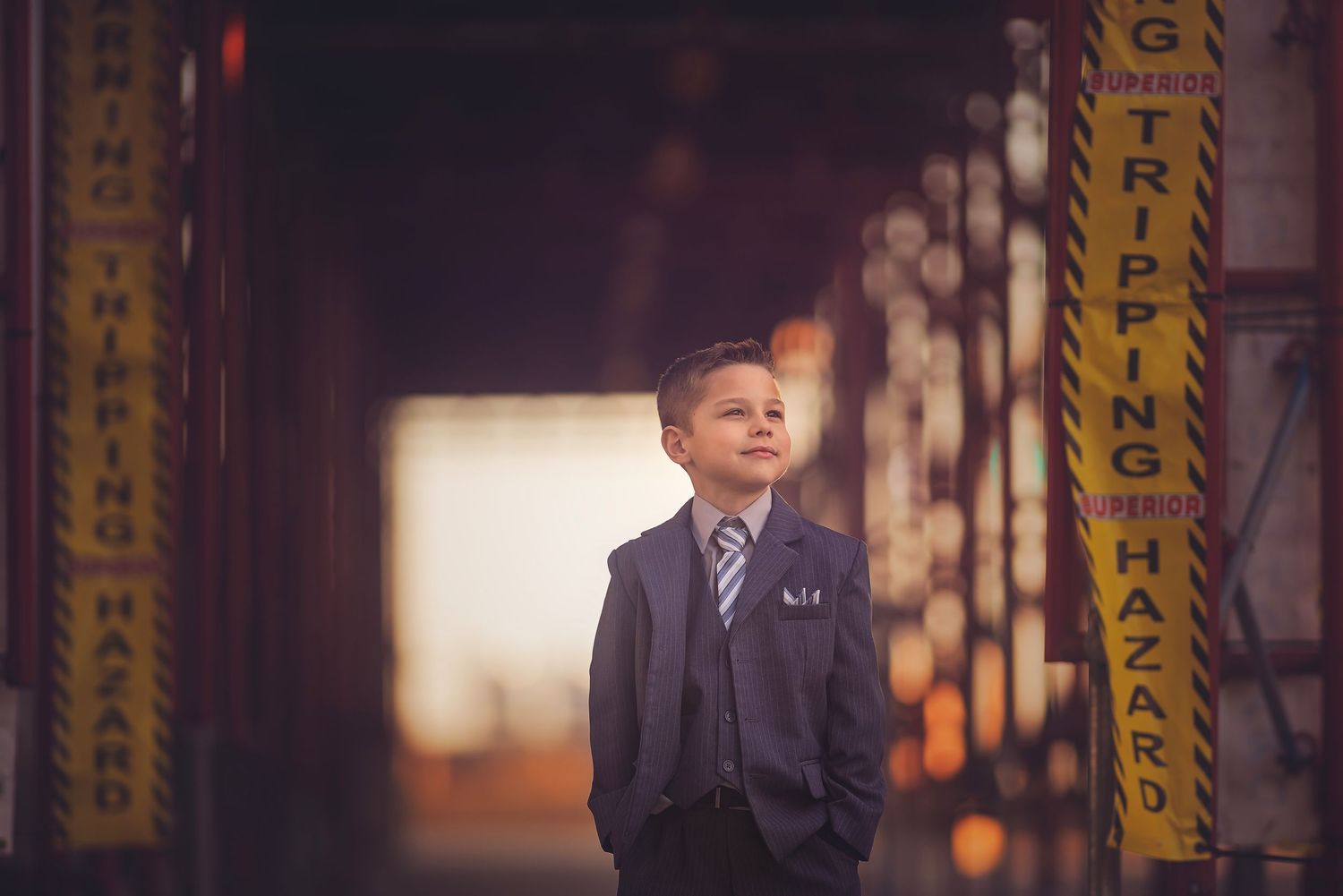 Young boy in a gray suit and striped tie stands in a warm-lit industrial corridor with yellow tripping hazard signs.