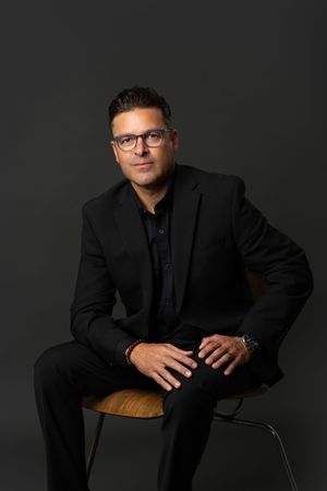 Man in glasses and an all-black suit sits on a wooden chair against a dark studio backdrop in a professional portrait.