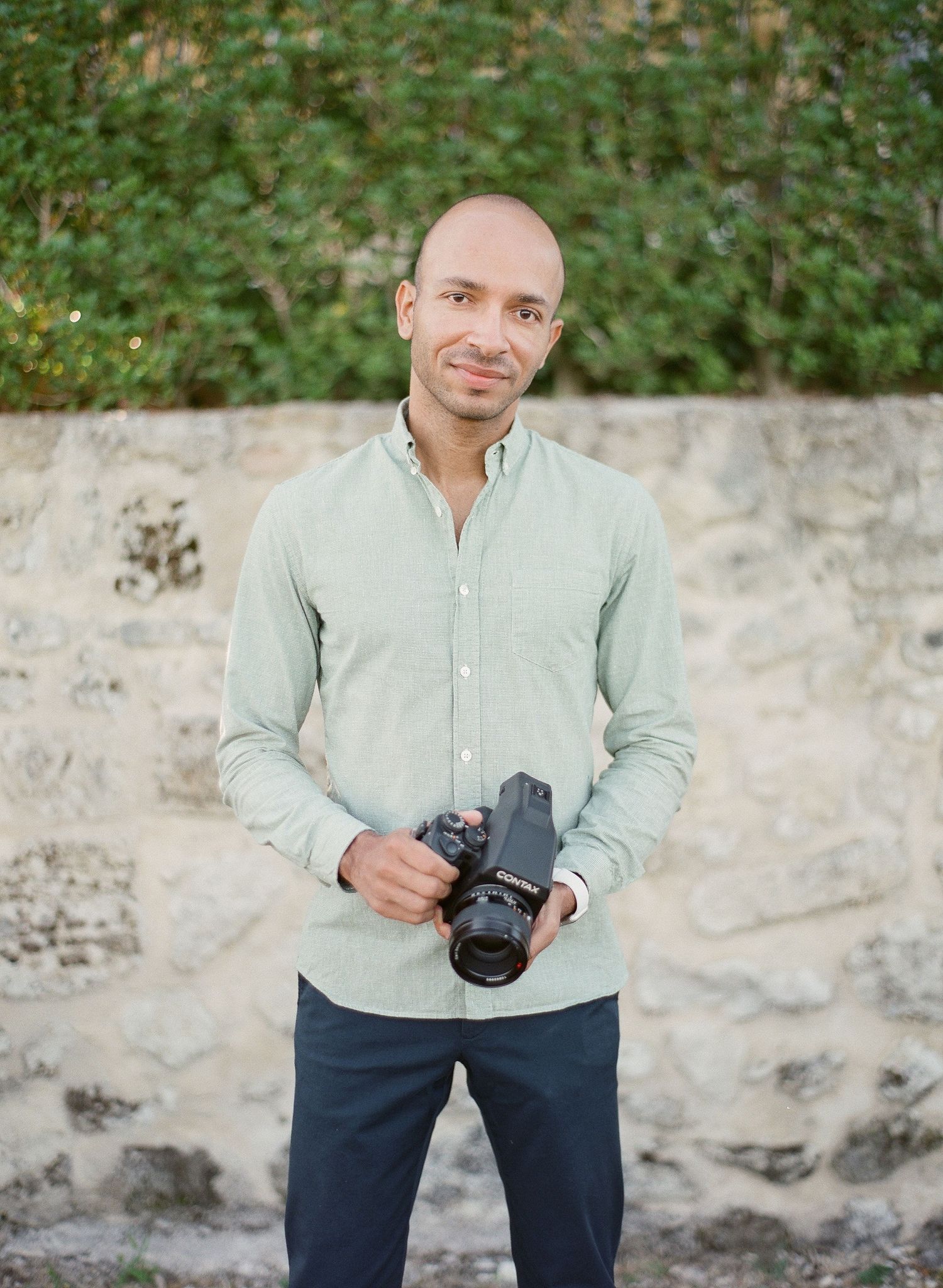 Un photographe professionnel en chemise boutonnée vert menthe tenant un appareil photo, avec un mur en pierre en arrière-plan.