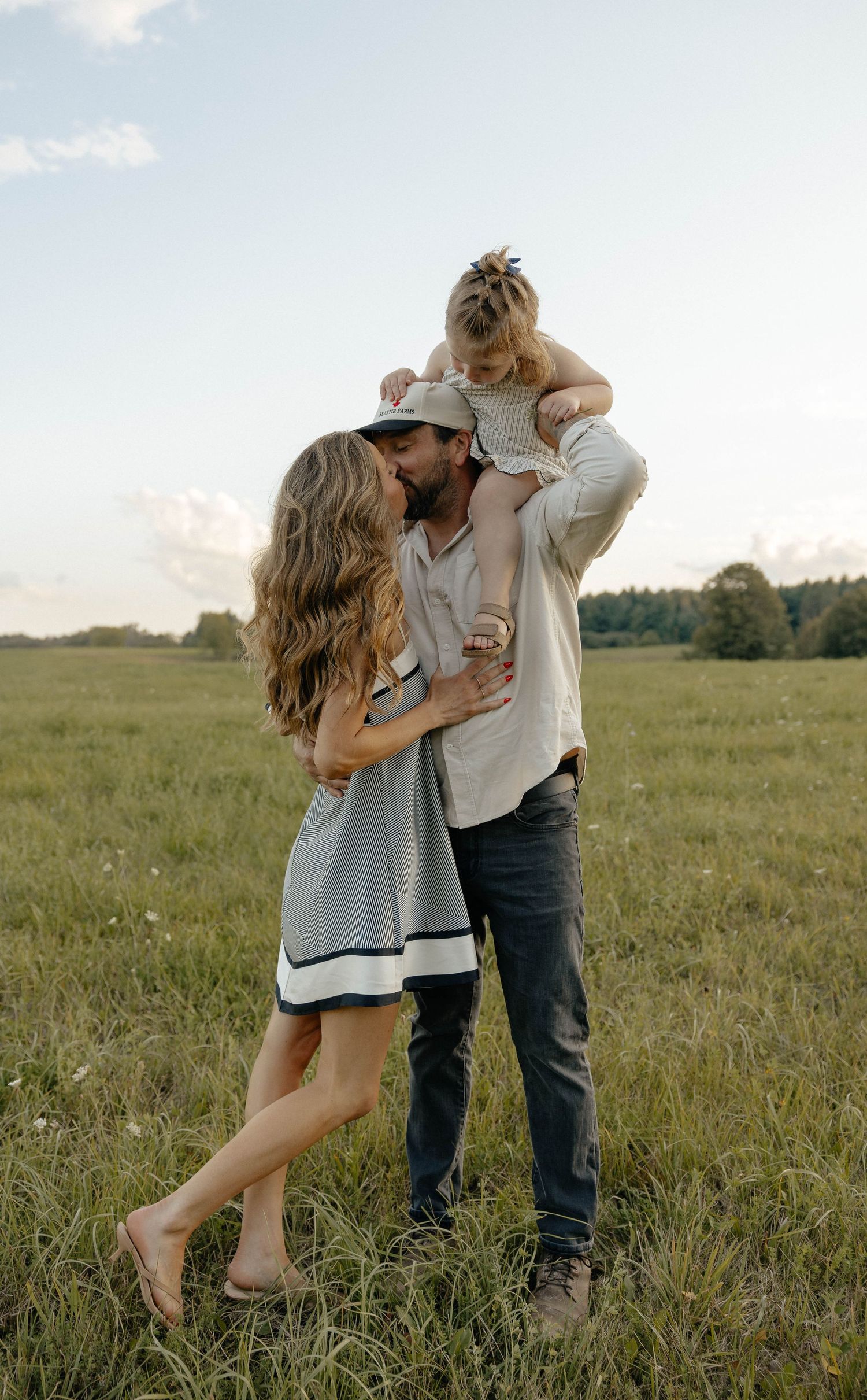 A couple shares a kiss in a green field while a young child sits on the man's shoulders during golden hour.