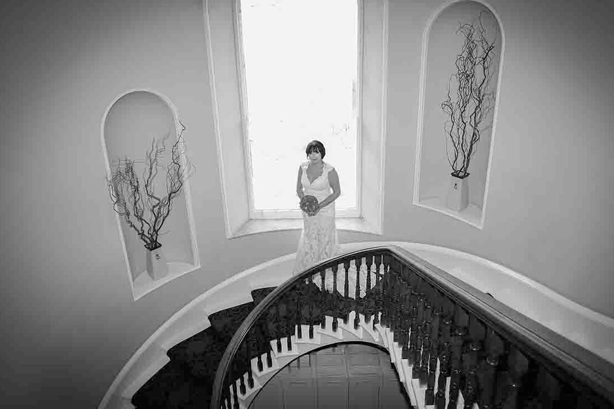 Black and white photo of a bride in a white gown standing at the top of an elegant curved staircase at Greenhill Hotel , Wigton, Cumbria