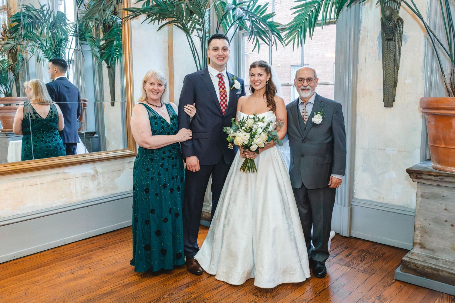 Wedding party of four posing indoors, bride in white gown holding bouquet, groom in suit, flanked by elegantly dressed family members.