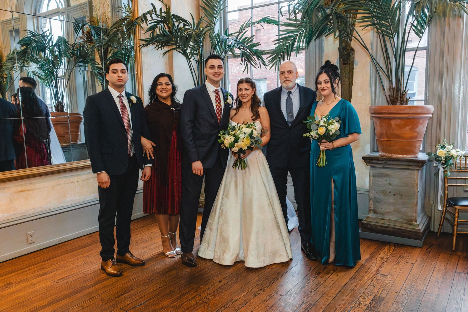 Wedding party poses indoors with bride in white gown, groom, and family members in formal attire near tropical plants.