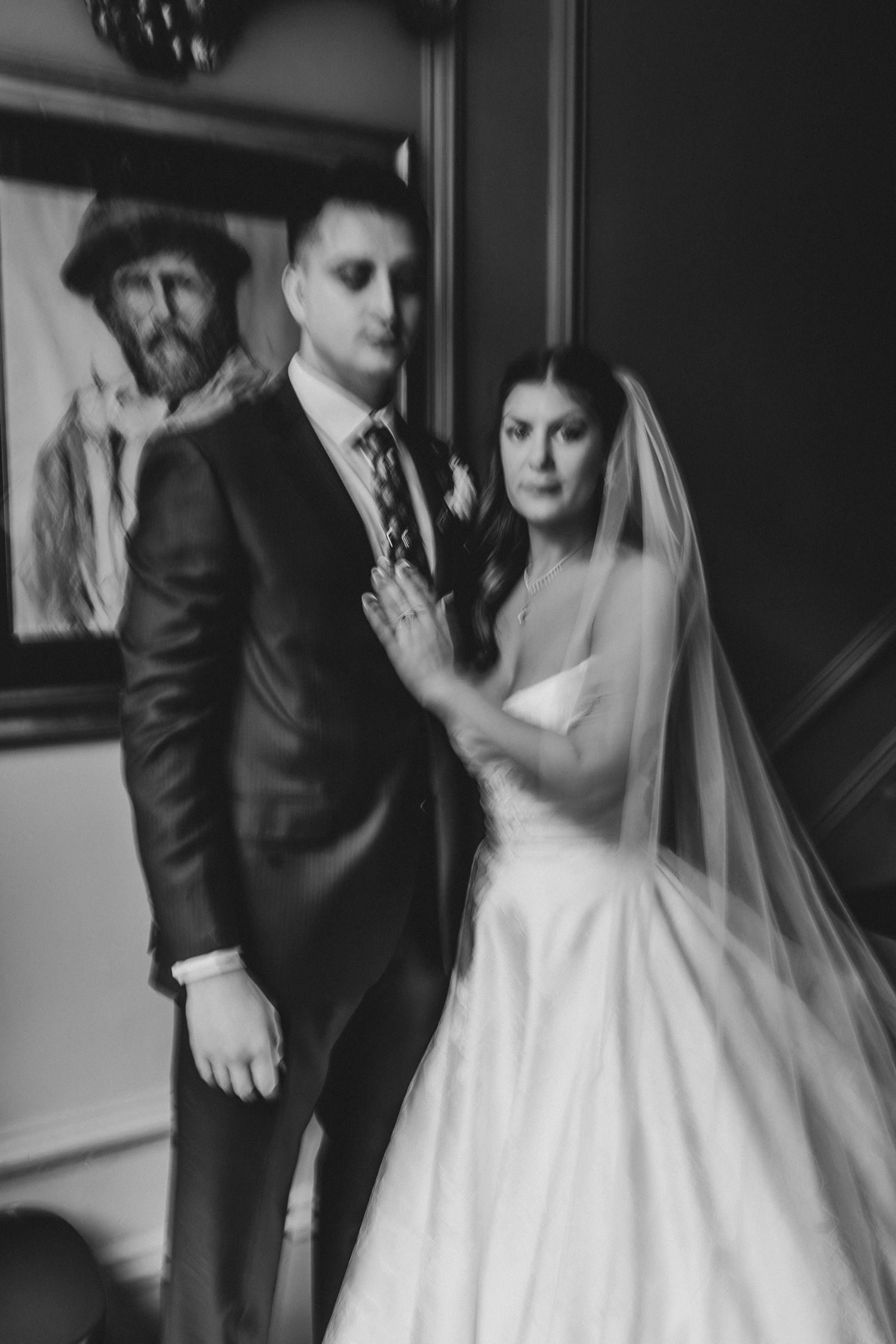 Black and white wedding photo of bride and groom standing together indoors, with a portrait painting visible in the background.
