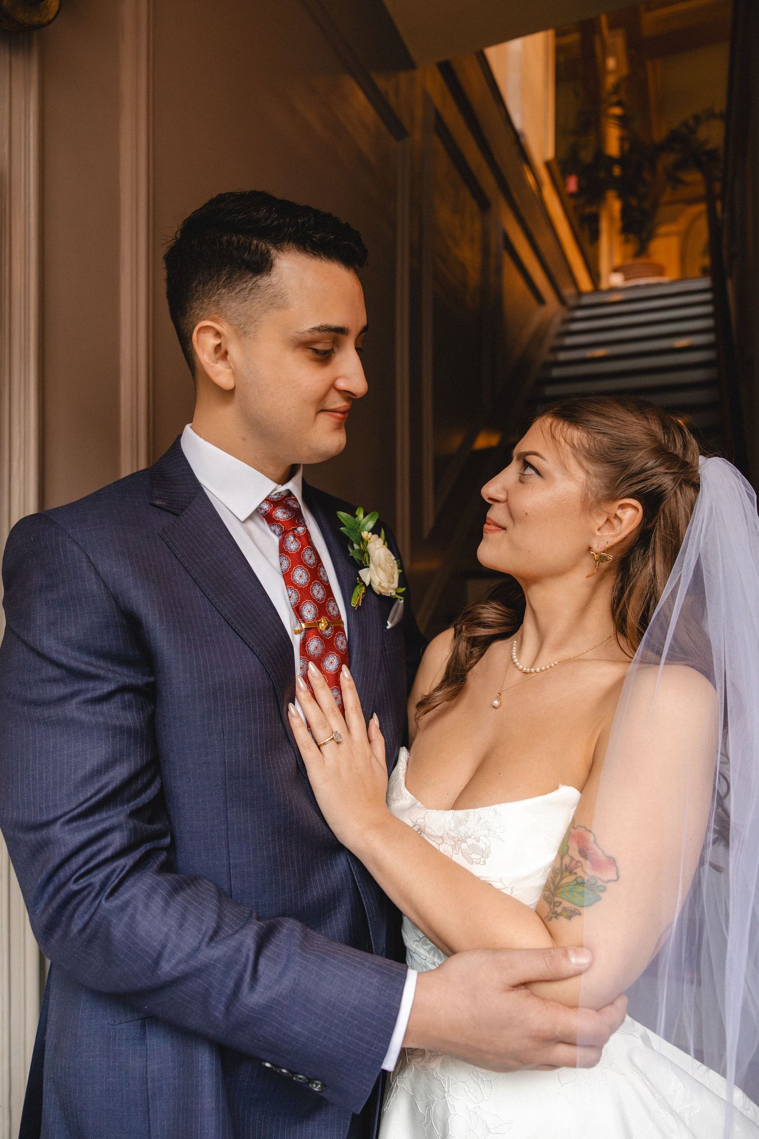 Bride and groom share a romantic moment, she in a white gown with veil, he in a navy suit with red tie.