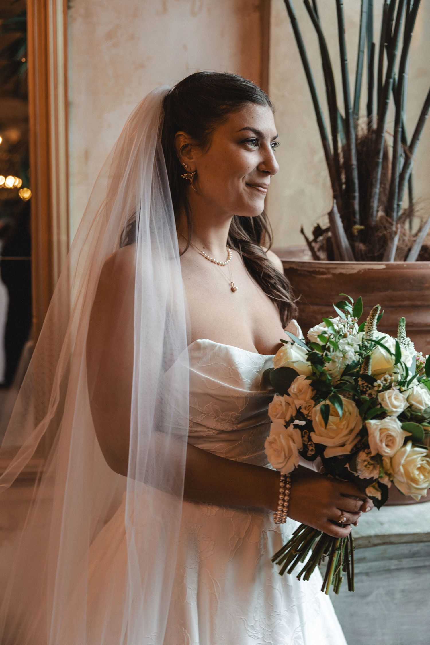 Smiling bride in white strapless gown and veil holds bouquet of cream roses, looking sideways in elegant venue.