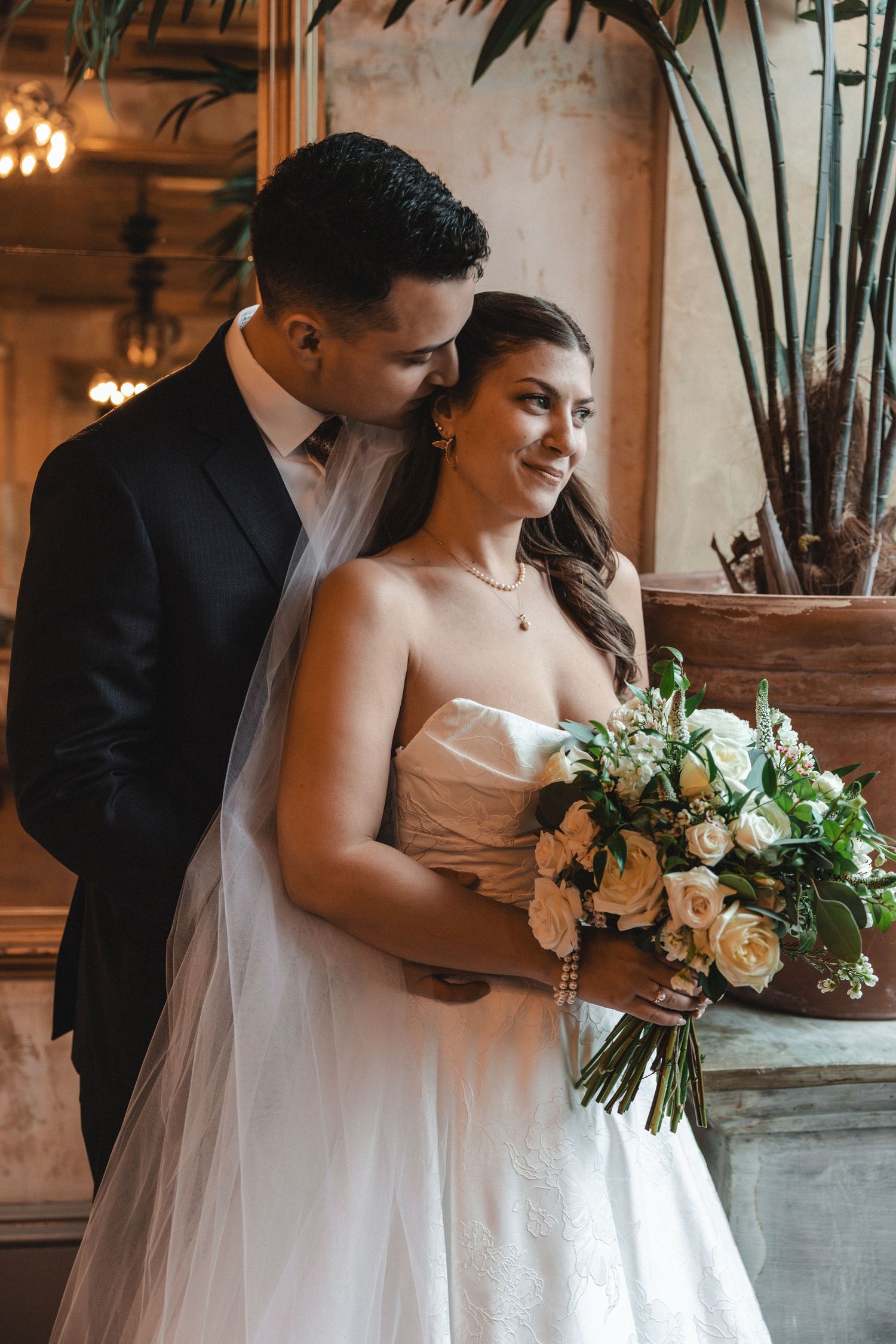 Bride and groom embrace indoors, bride holding white rose bouquet in elegant strapless gown, groom in black suit.