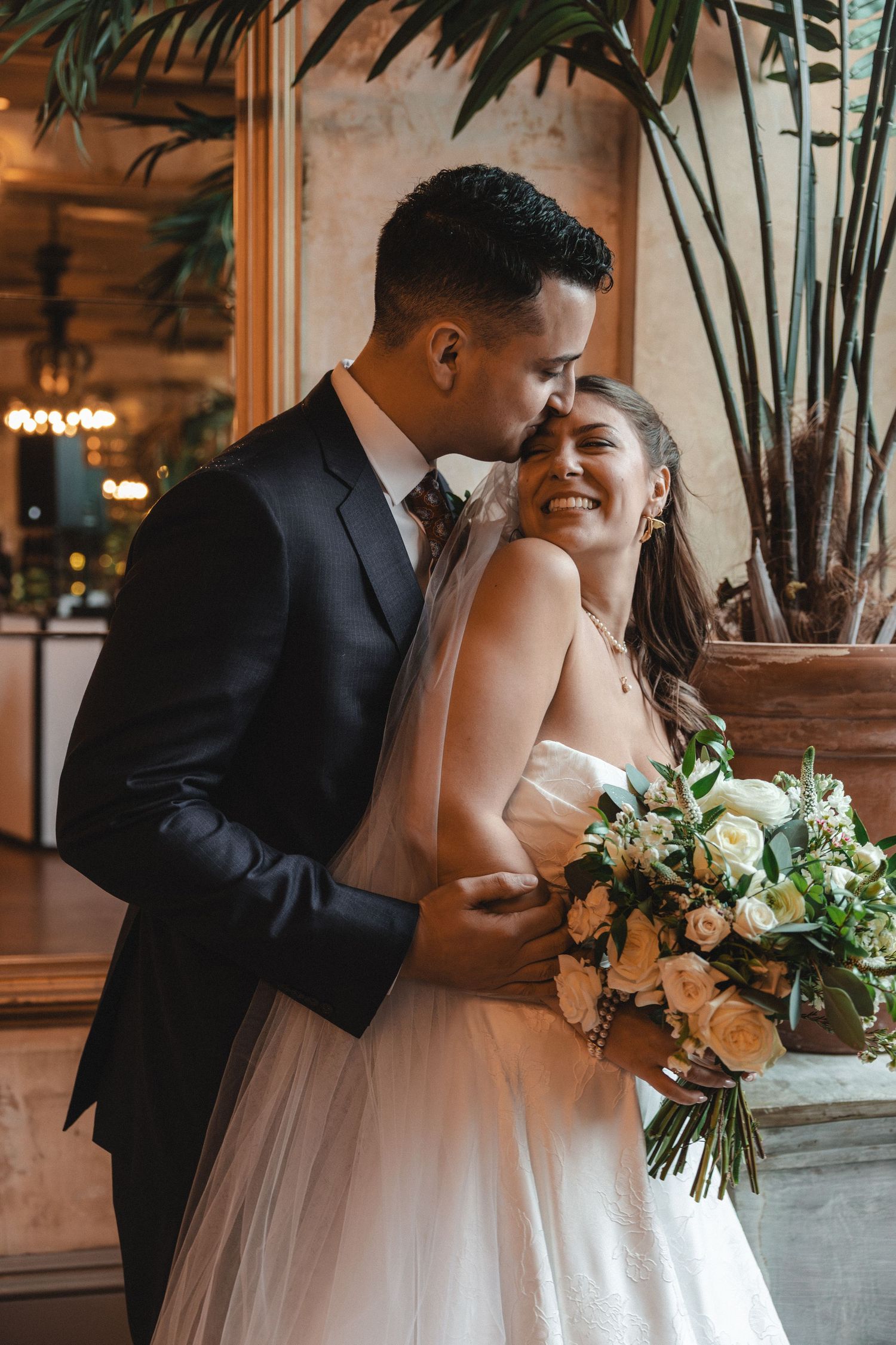 Groom in black suit kisses bride wearing white gown holding bouquet of white and peach roses indoors.