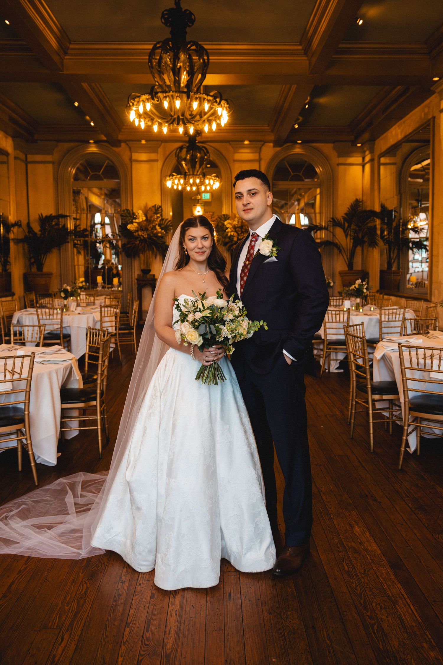 Bride and groom pose in a grand ballroom with chandeliers, bride in a strapless gown holding a white bouquet.