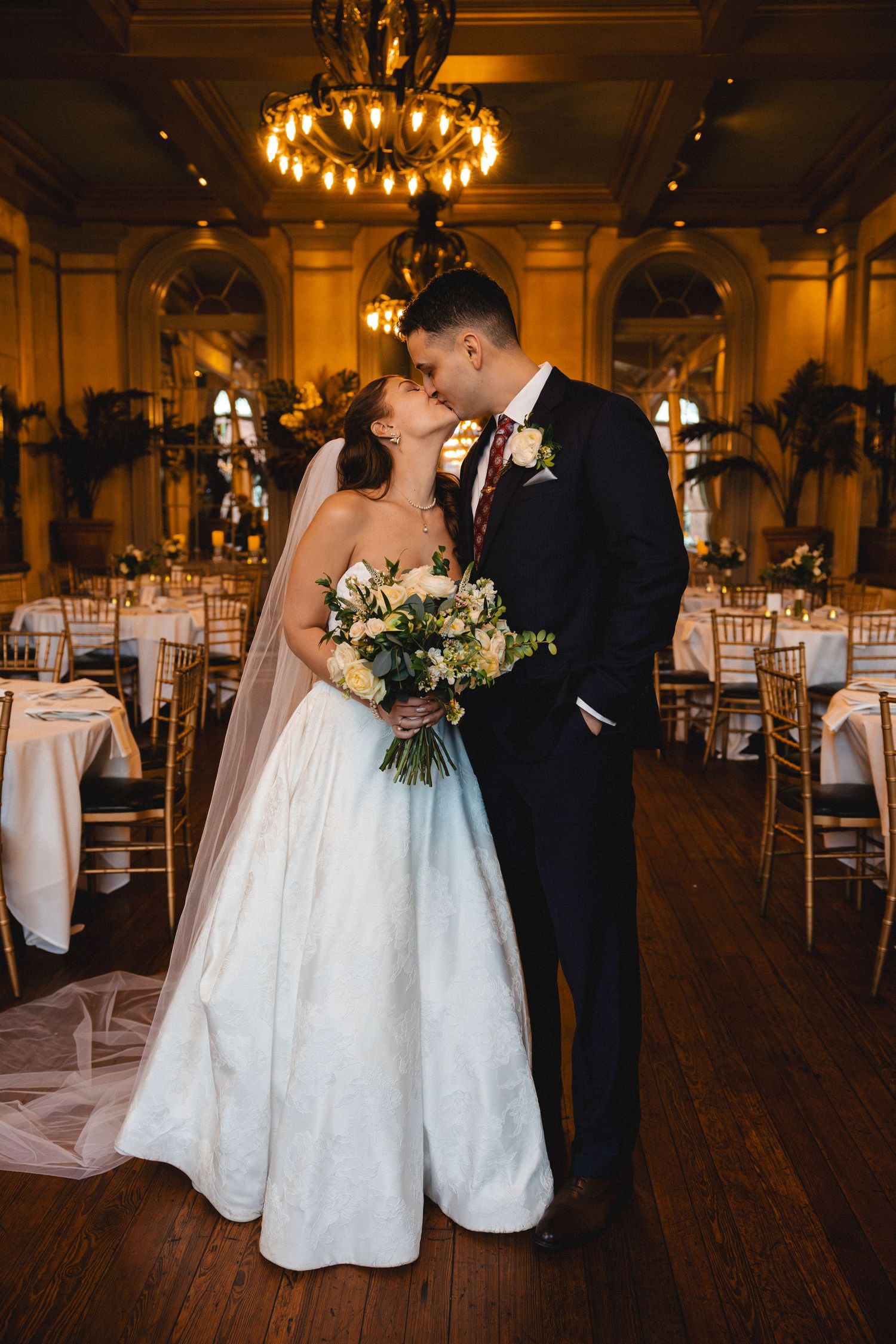 Bride and groom share a romantic kiss in an elegant ballroom, bride holding a white floral bouquet wearing a veil.