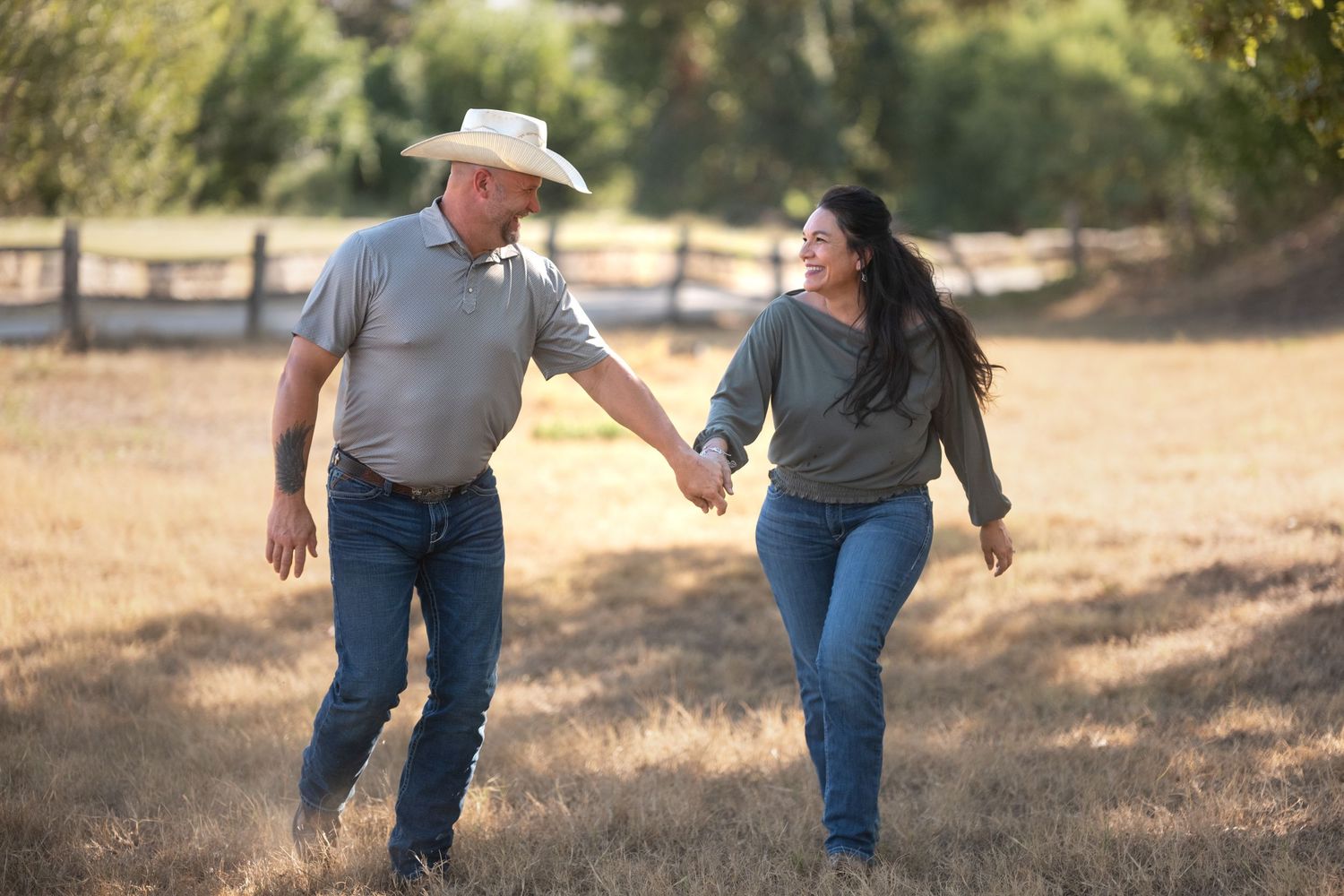 Couple holding hands and walking through a sunny rural field, man wearing cowboy hat and woman with long dark hair.