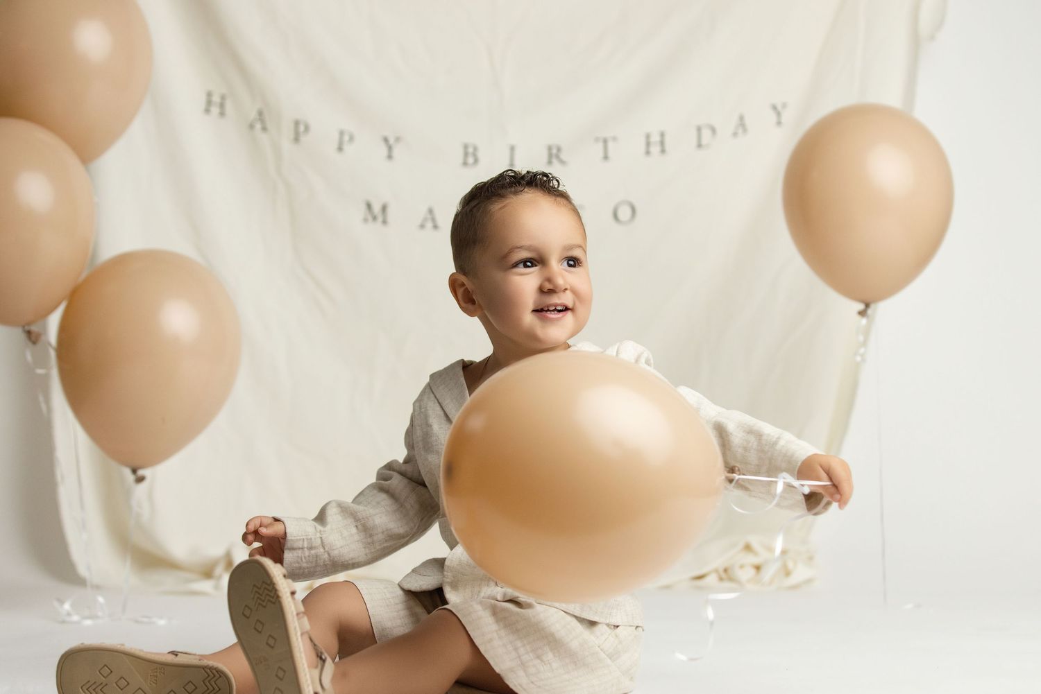 A smiling toddler sits holding a large beige balloon in front of a Happy Birthday banner with matching neutral balloons.