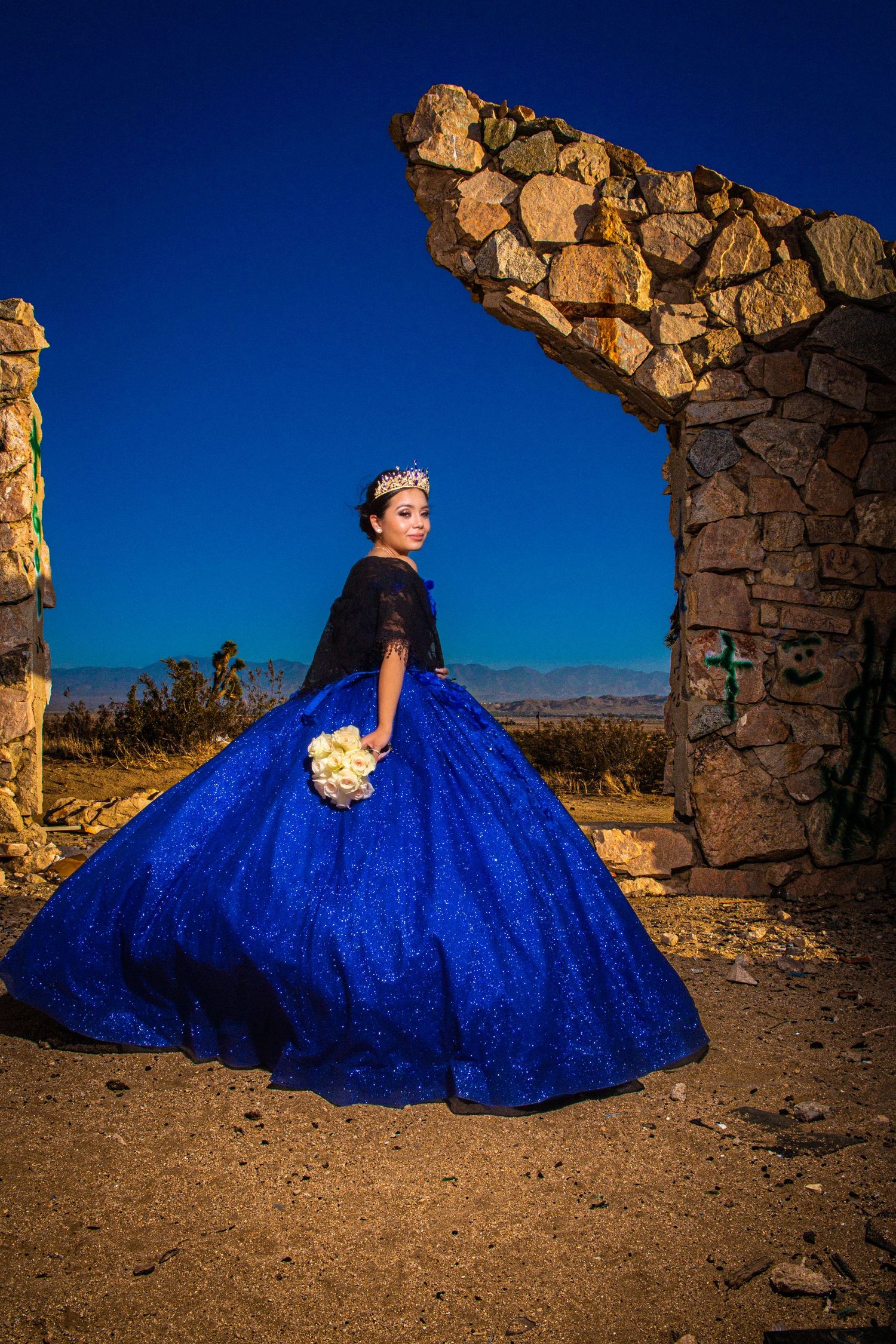 A person in a vibrant royal blue sequined ballgown poses by stone ruins at sunset in the desert.
