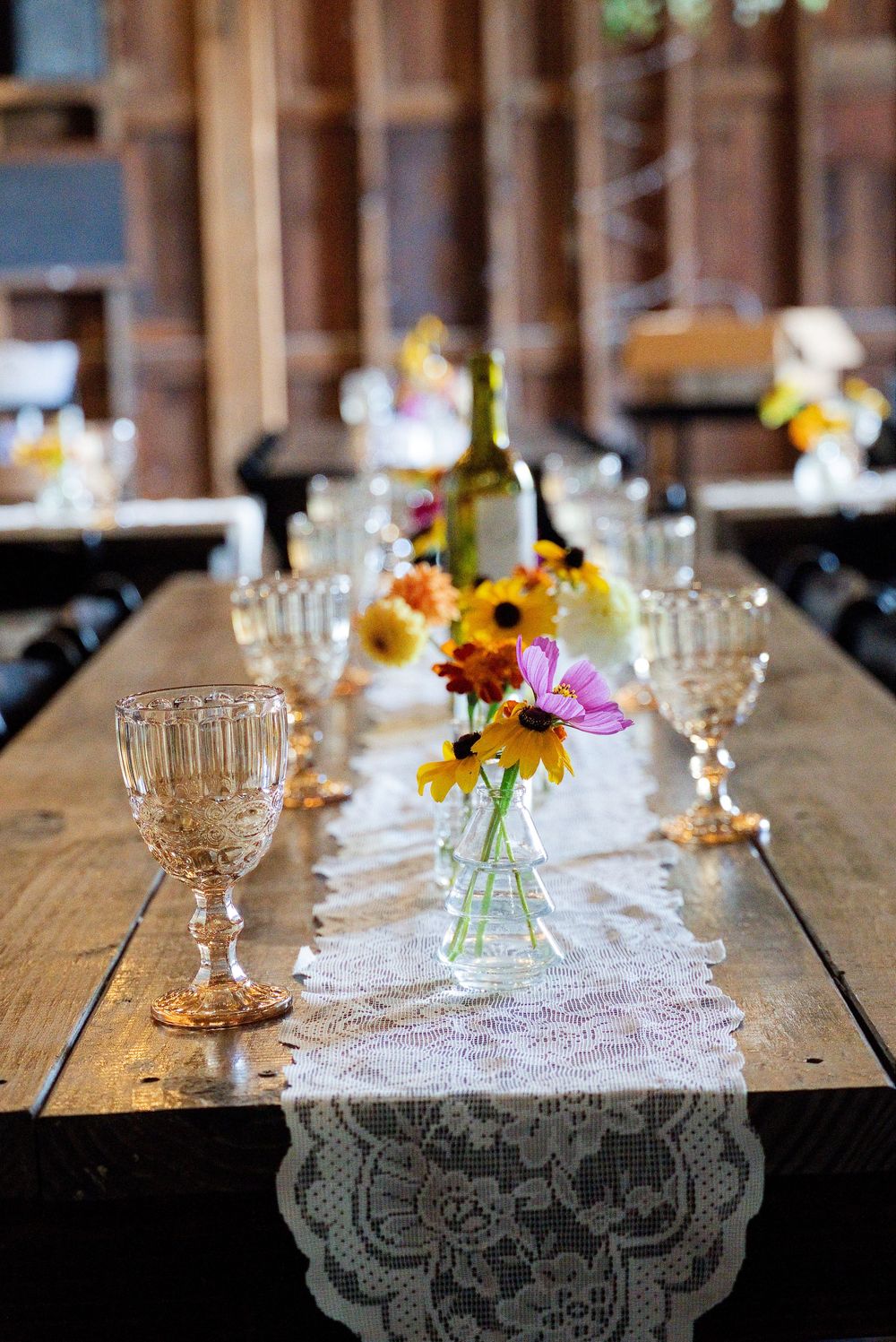 Rustic wedding reception tables at The Centennial Barn in Wayland, MI with lace runners, vintage pink glassware, and wildflowers.
