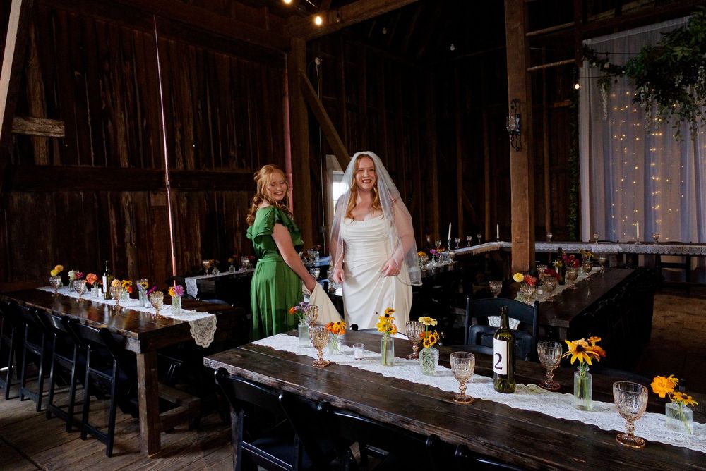 Bride in a classic gown with romantic curls stands by her maid of honor in a green dress inside the barn reception area.