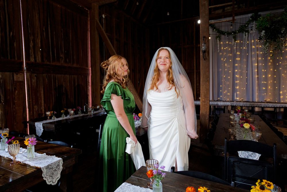 A bride in a classic wedding gown and veil stands with her maid of honor in a green dress inside the reception area at The Centennial Barn.