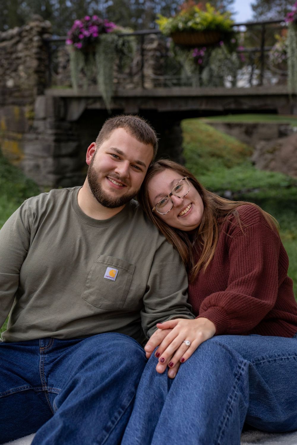 Engaged couple smiling in front of scenic bridge with flowers