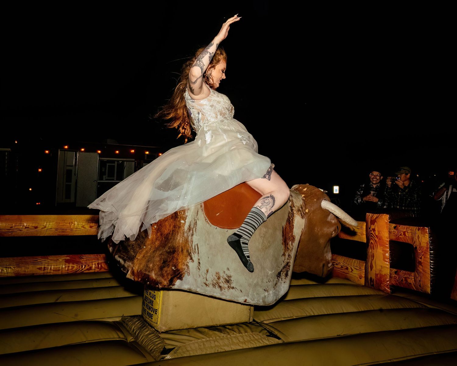 Girl in white dress riding mechanical bull at night outdoor event, wearing striped socks, arms raised.