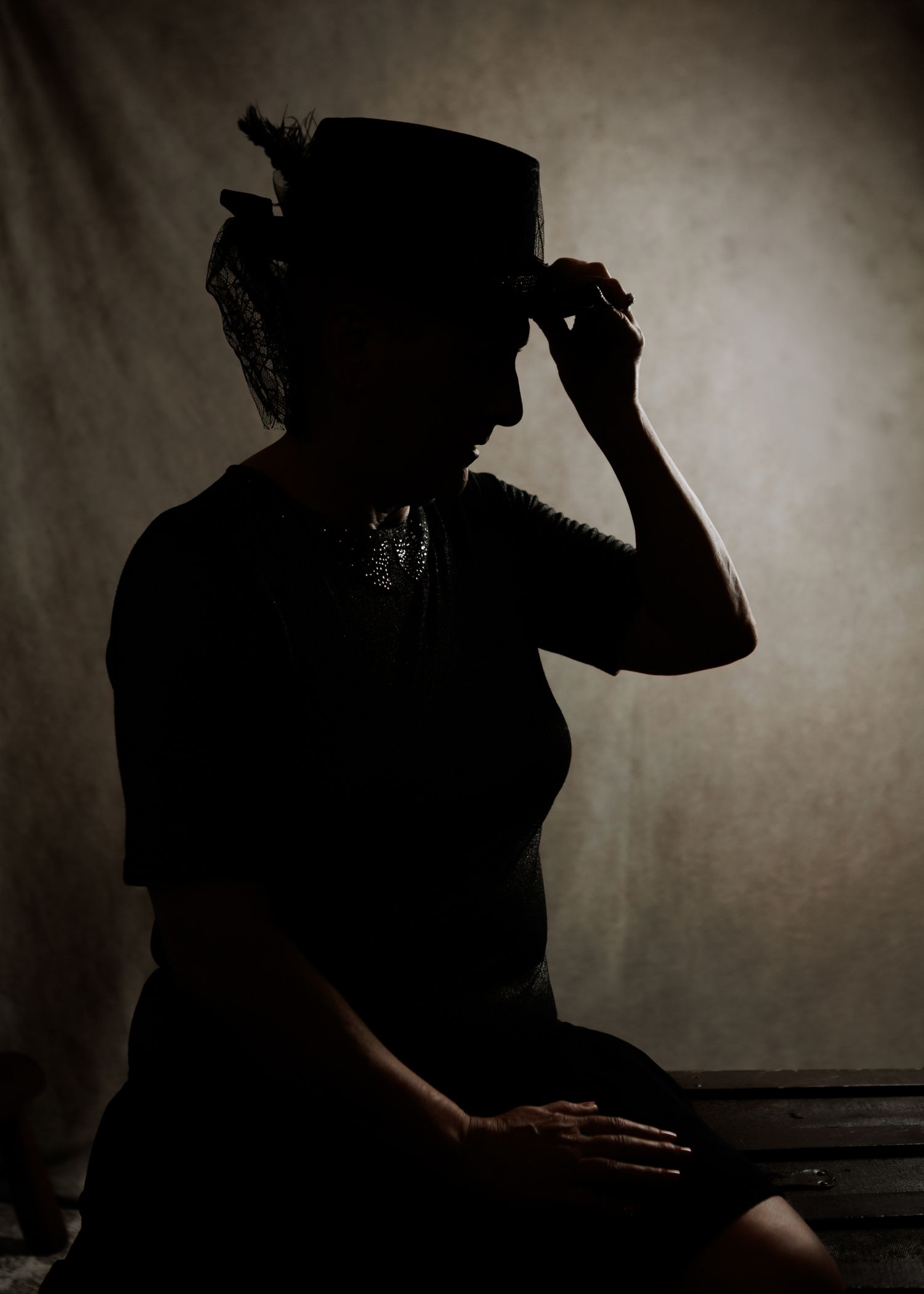 Silhouette of a woman in a dark outfit touching her decorative hat against a moody studio backdrop almost revealin the effects of hair loss.