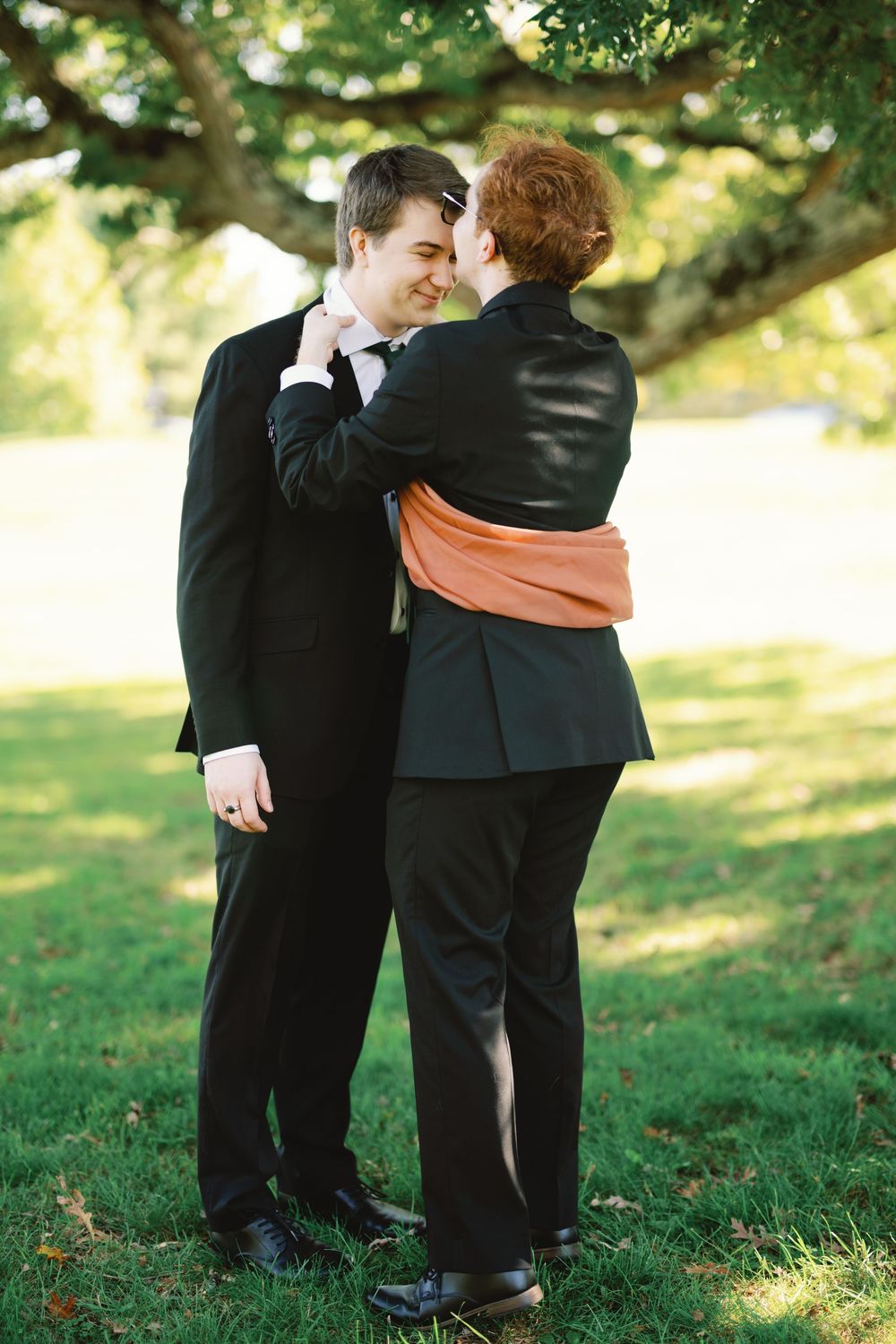 Couple in formal black attire sharing a romantic moment under a large tree on a green lawn.