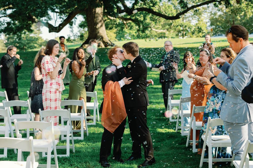 Two brides share a kiss while guests toss confetti during an outdoor wedding ceremony on a lush green lawn.