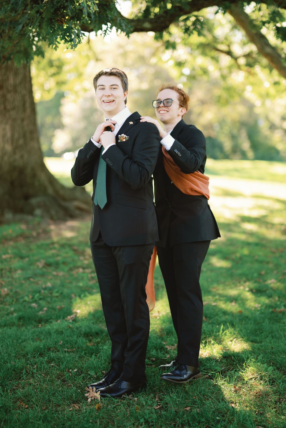 Two young men in black suits pose outdoors under a tree, one adjusting his green tie and the other wearing sunglasses.