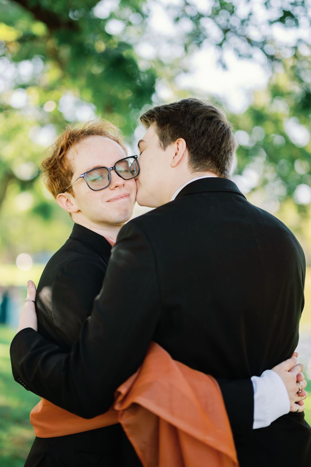 Two men in black suits share a tender embrace and kiss outdoors, surrounded by lush green trees.