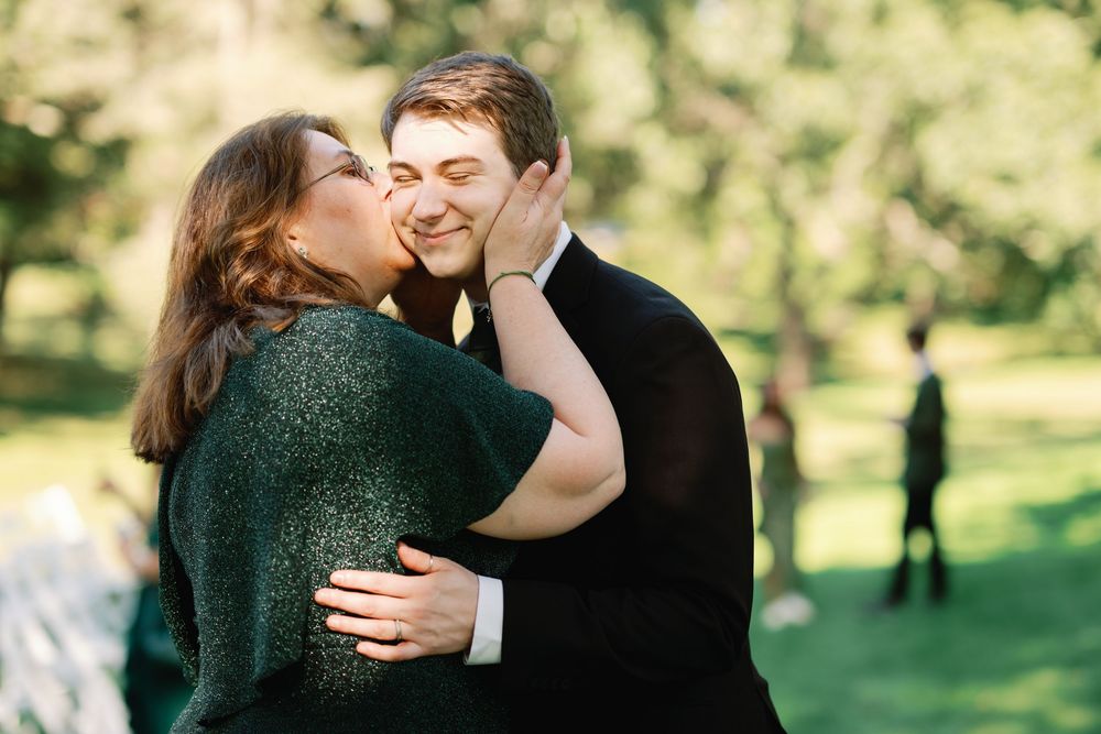 A couple shares a tender kiss outdoors at a garden wedding ceremony, surrounded by lush green trees.