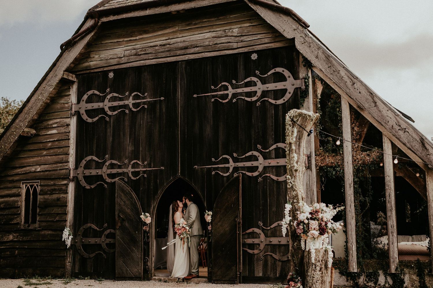 Couple poses before rustic wooden barn with decorative iron hardware at outdoor wedding venue.