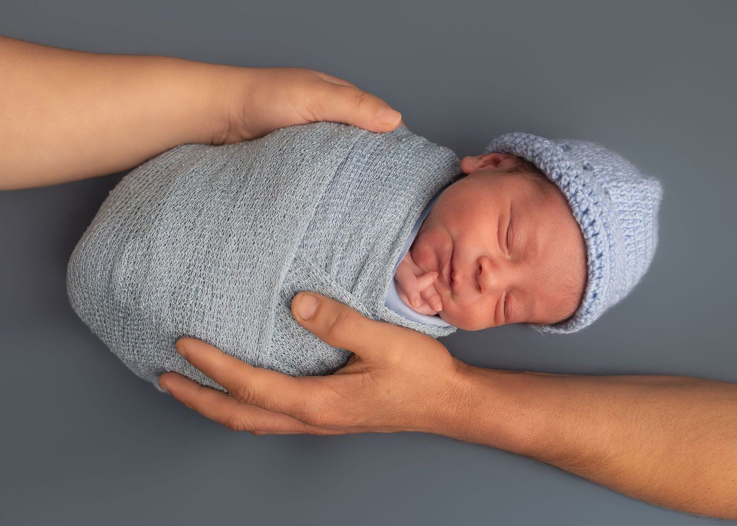 Newborn baby swaddled in a soft gray knit wrap and matching hat, cradled gently in adult hands.