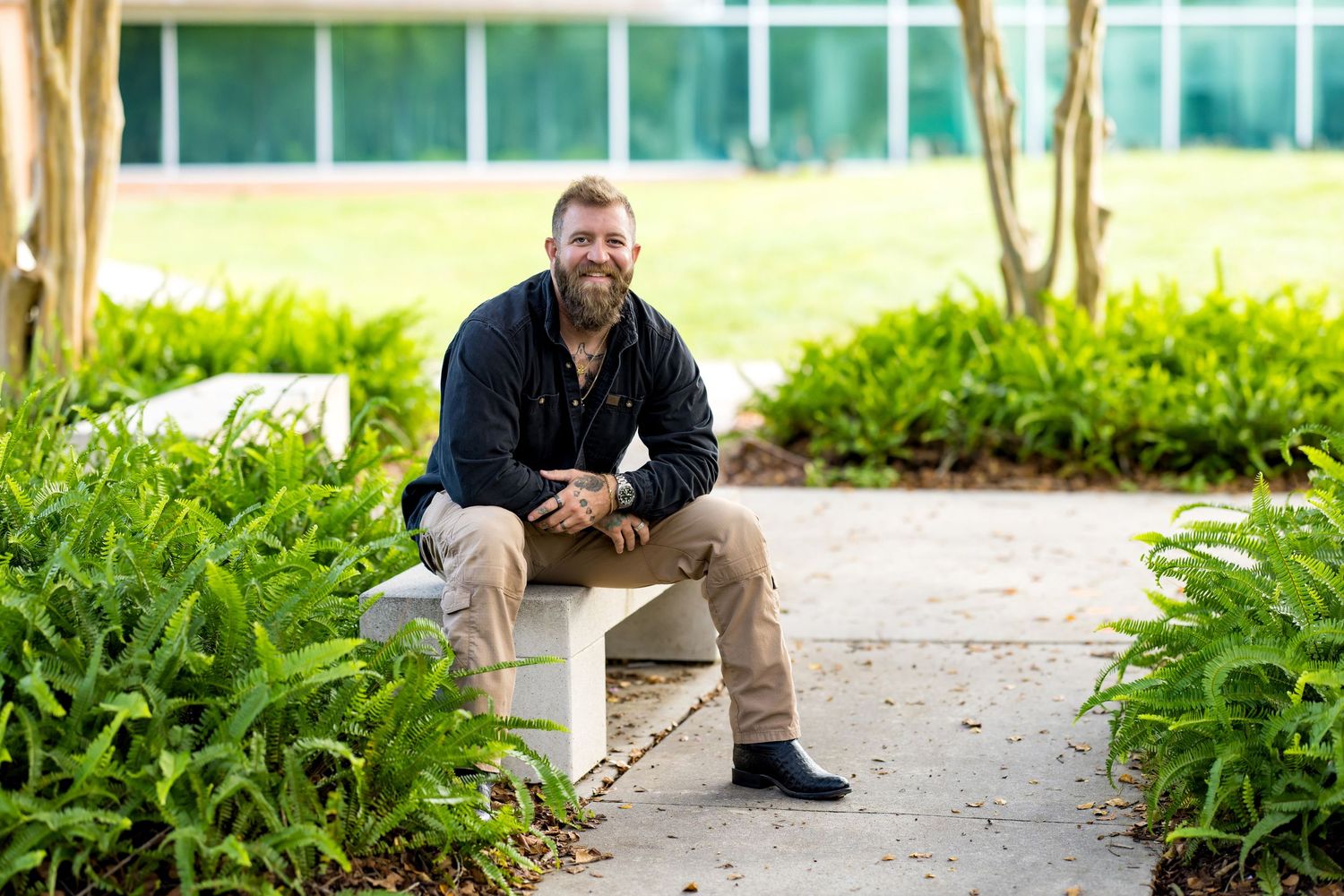 Bearded man sitting on a white bench surrounded by lush green plants in an outdoor garden setting.