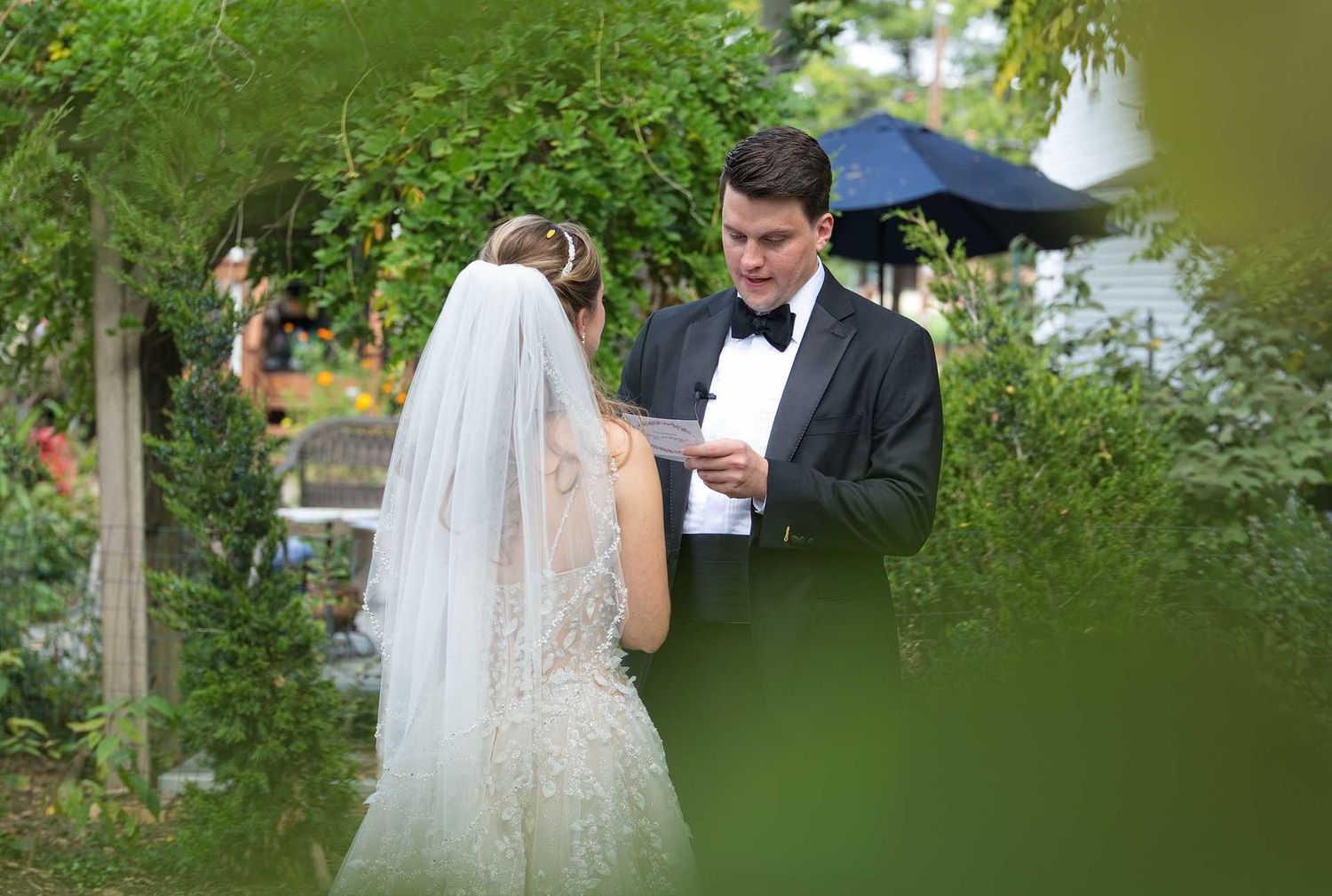 Groom reads wedding vows to bride in white gown and veil during outdoor garden ceremony.