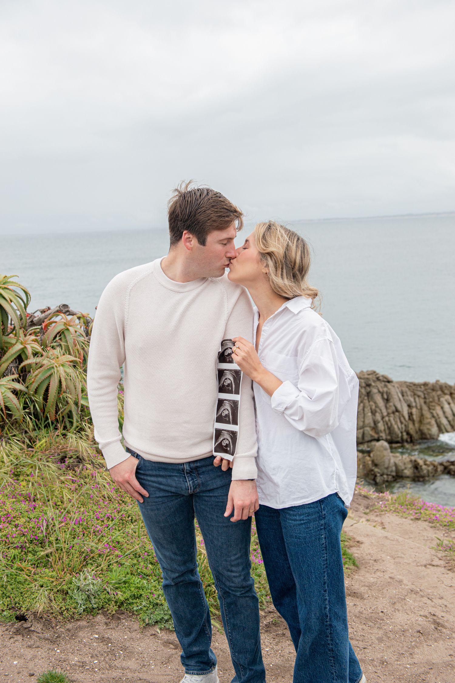 Couple kissing outdoors by ocean cliffs, holding ultrasound photos, announcing pregnancy in a coastal setting.