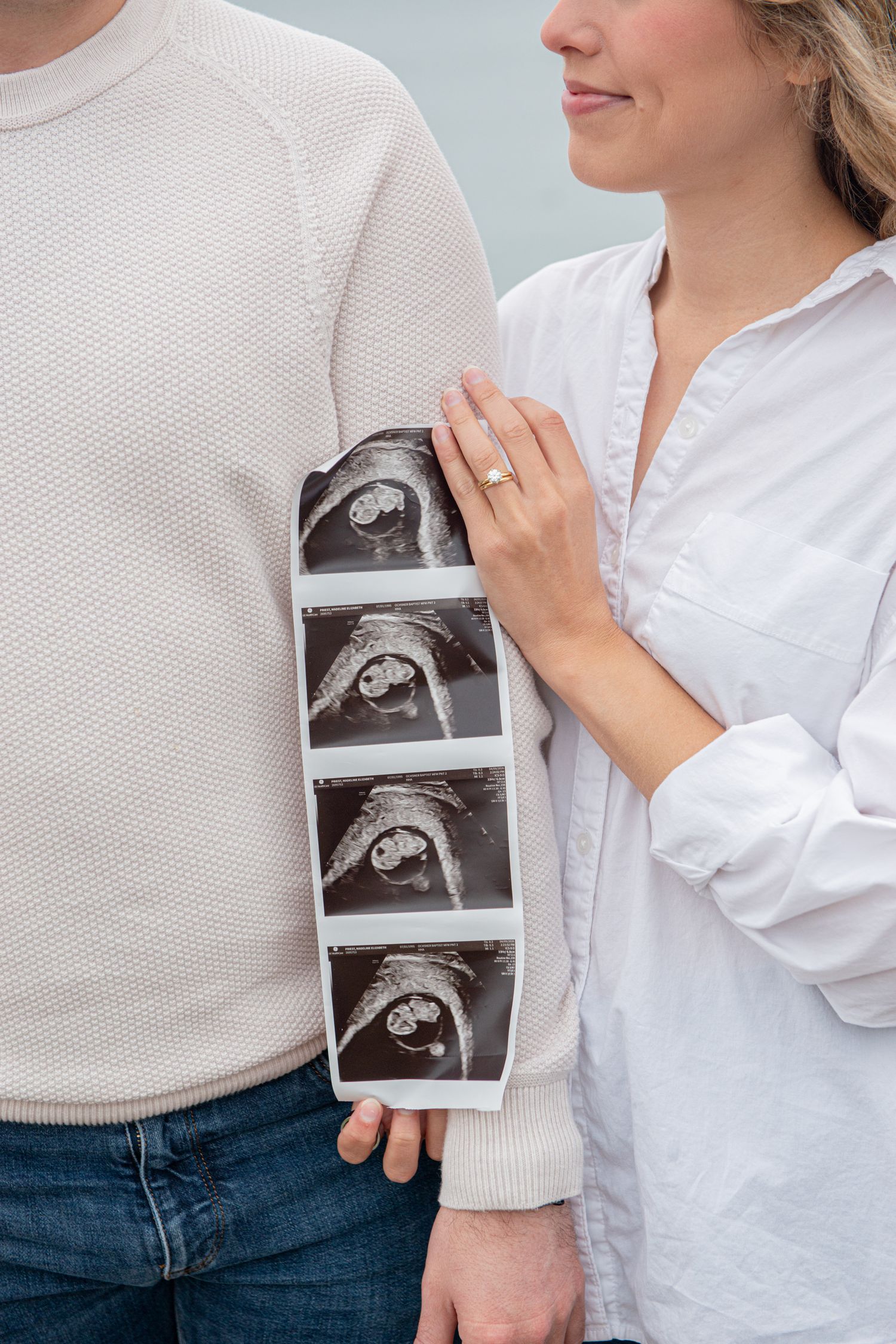 Couple holding ultrasound photos, woman in white shirt with engagement ring smiling, pregnancy announcement photo.