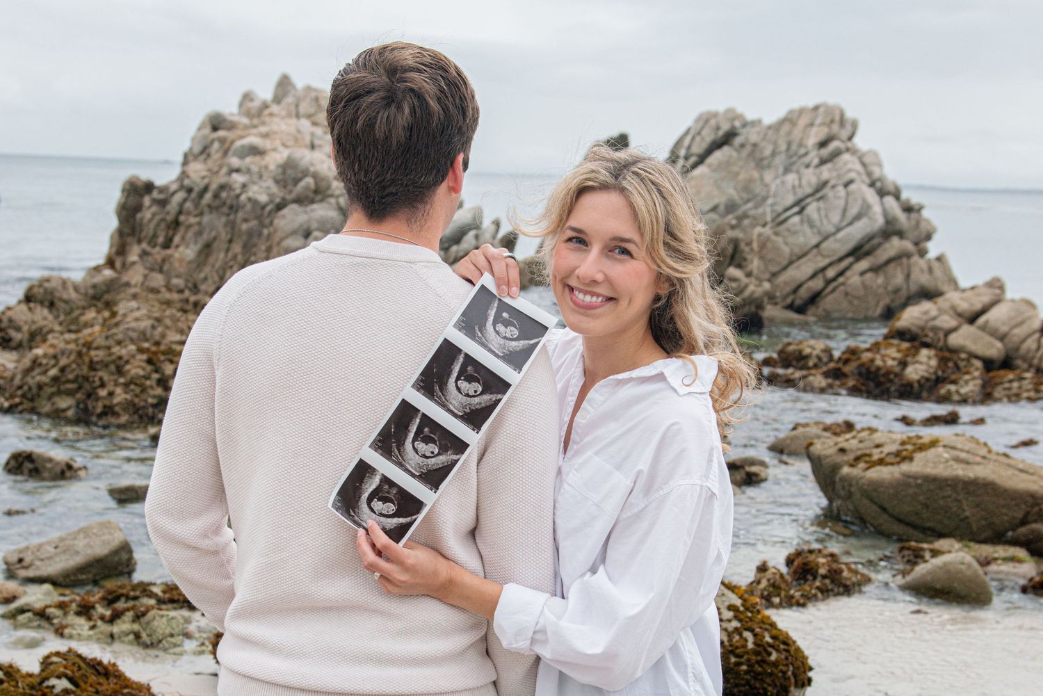 Couple at rocky beach holding ultrasound photos, woman smiling at camera in pregnancy announcement photo.